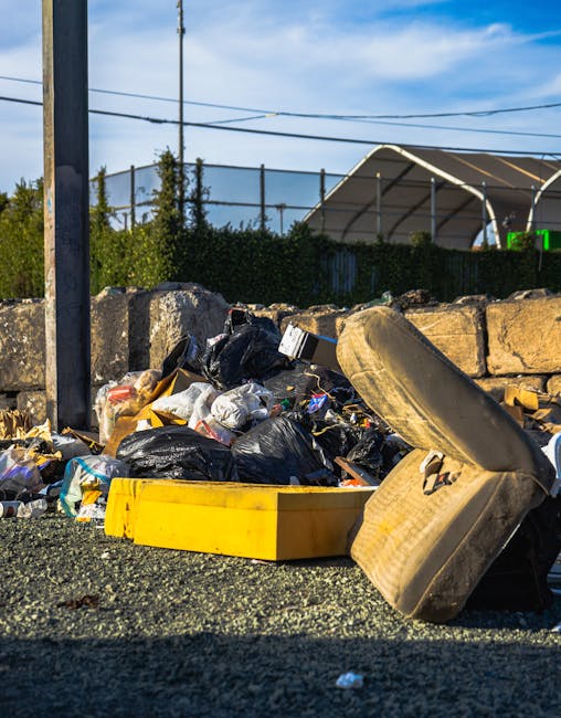 A pile of mixed waste materials is scattered on the ground in an outdoor setting, including several black plastic rubbish bags filled with debris, an empty yellow plastic container, an overturned worn beige car tire, and assorted smaller debris such as paper and plastic wrappers. The scene is situated on a paved surface with a low stone wall behind the rubbish, composed of rough-textured brown blocks. In the background, a metal fence surrounds what appears to be a sports facility or industrial area, with green hedges and a partially visible white tent-like structure. A utility pole with wires is seen to the left, and the sky above is partly cloudy with patches of bright blue, suggesting daytime. This scene reflects a typical example of unmanaged refuse that a professional rubbish removal service such as Rubbish Removal Islington might collect as part of an independent waste clearance operation, emphasizing the importance of proper disposal of household or commercial waste to maintain a safe environment.
