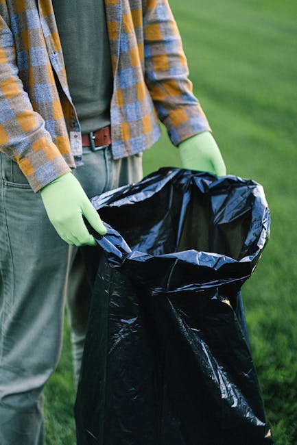 The image depicts a person wearing a checked yellow and blue jacket, green gloves, and grey trousers, holding a large black rubbish sack open with both hands. The sack, made of plastic with a shiny, crinkled texture, is positioned in an outdoor setting on a grassy area. The background features a blurred expanse of well-maintained green grass, suggesting the scene is taking place in a garden or park. The individual's torso and hands are visible, while their face is not shown. The setting indicates an activity related to waste collection or rubbish disposal away from municipal waste services, possibly part of a private or independent rubbish removal effort. The scene emphasizes proper handling and containment of waste in preparation for disposal or collection, aligning with professional rubbish removal practices. The lighting appears natural, with soft, even illumination highlighting the textures of the plastic sack, gloves, and clothing, reflecting an environment suitable for on-site waste clearance activities as conducted by companies like Rubbish Removal Islington.