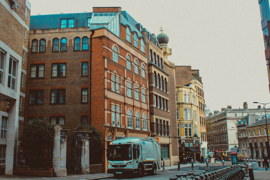 A daytime city street scene featuring a row of historic multi-storey buildings with brick and stone facades, large arched and rectangular sash windows, and ornate architectural details. A modern street-cleaning vehicle, painted in light blue with the brand name 'DENNIS' visible on the front, is parked on the pavement near the entrance of a gated property with stone pillars and iron fencing. The street is lined with bollards, and behind the vehicle, a line of bicycles is secured to a bike rack. In the background, other buildings display classical and Victorian architectural elements, including decorative cornices and windows, with a pale yellow building featuring bay windows and a domed rooftop structure. The scene is illuminated by natural daylight, with a slightly overcast sky, and the overall atmosphere is of an urban environment that could be suitable for private waste collection or rubbish removal services, as represented by the presence of the waste collection vehicle from Rubbish Removal Islington, situated within the context of alternative waste handling methods outside local authority collection.