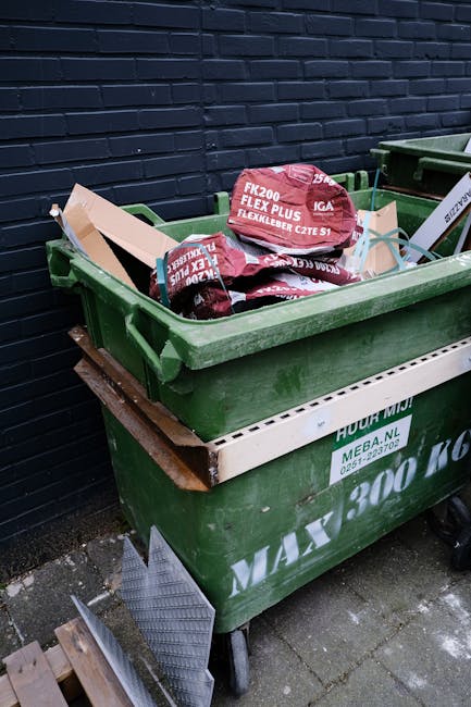 A green wheeled rubbish container filled with red and black bags of garden waste or waste bags resting on a white plastic crate, positioned against a dark brick wall outdoors. The container is situated on a paved surface, with a small wooden pallet underneath part of it. The bags appear to contain materials such as soil, leaves, or garden debris, and the scene suggests an on-site waste collection or private disposal method. The environment indicates a residential or commercial area where rubbish removal services like those provided by Rubbish Removal Islington might handle such waste. The overall setting highlights a typical outside waste storage area, with minimal natural light illuminating the scene and the container's environment in a neutral, practical context for waste management services.