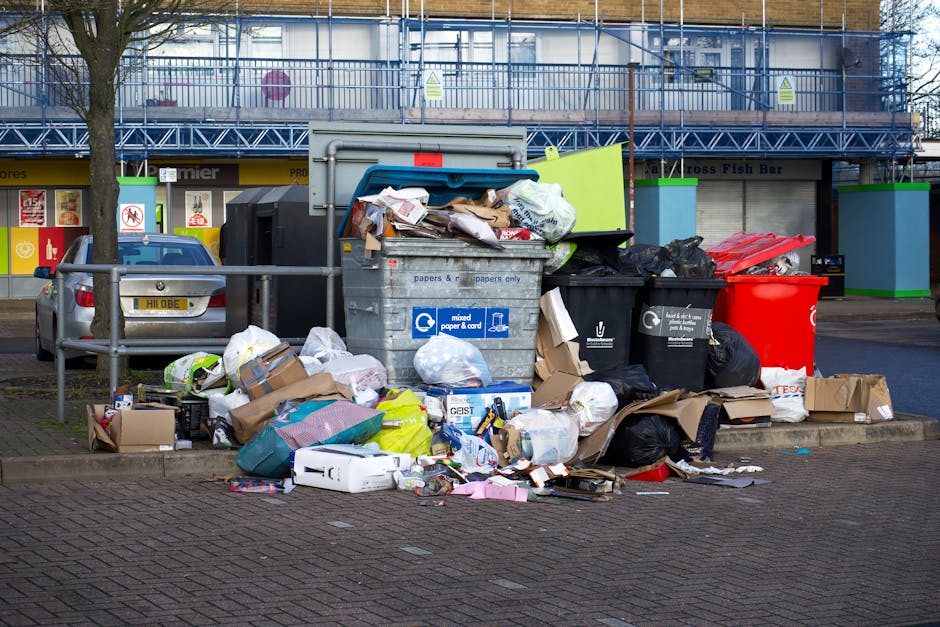 A large accumulation of mixed household waste and recycling including cardboard boxes, plastic bags, paper, and electronic packaging piled around a grey communal bin marked for mixed paper and card in an outdoor urban setting. The trash extends onto the nearby pavement, with some items spilling onto the road. Several black and red trash bags are placed next to the bin, some partially opened, revealing more waste inside. Behind the pile, a parked silver car is visible, along with a metal railing separating the waste area from the vehicle. In the background, there is a building under renovation with scaffolding and blue safety netting, and a row of storefronts with yellow signage indicating commercial premises. The scene is lit by natural daylight, and the environmental context suggests an area where private or independent waste disposal services, such as rubbish removal, might be required to manage the overflowing rubbish collection.