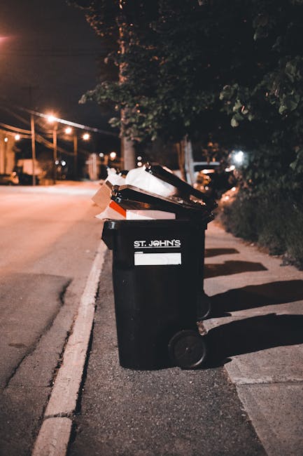 A green wheeled rubbish container filled with red and black bags of garden waste or waste bags resting on a white plastic crate, positioned against a dark brick wall outdoors. The container is situated on a paved surface, with a small wooden pallet underneath part of it. The bags appear to contain materials such as soil, leaves, or garden debris, and the scene suggests an on-site waste collection or private disposal method. The environment indicates a residential or commercial area where rubbish removal services like those provided by Rubbish Removal Islington might handle such waste. The overall setting highlights a typical outside waste storage area, with minimal natural light illuminating the scene and the container's environment in a neutral, practical context for waste management services.