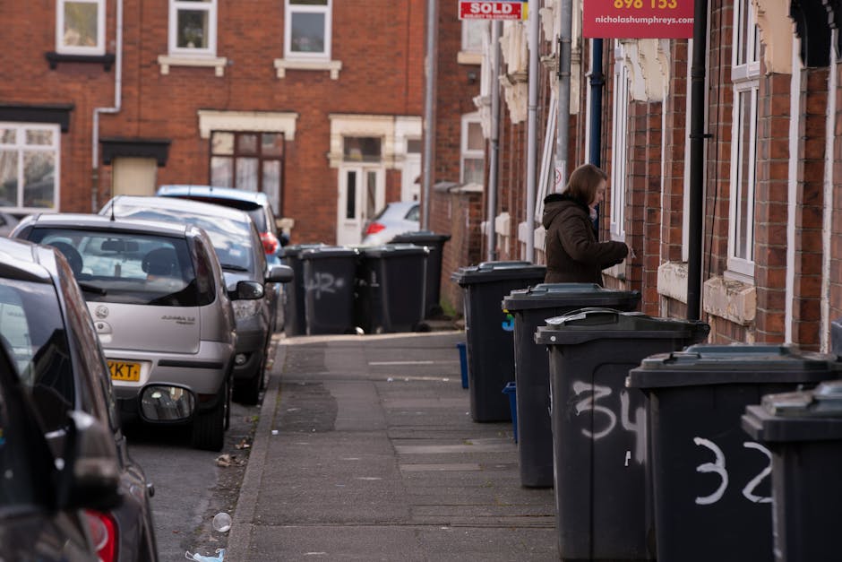 A residential street scene in Islington featuring a row of dark grey wheelie bins with white painted numbers on their sides, lined up along the sidewalk close to a brick building with white window frames. A woman dressed in a brown coat is opening or retrieving items from one of the bins on the right side of the image. Several parked cars, including a silver hatchback closest to the foreground and others behind it, are situated along the street. The pavement appears slightly worn with scattered litter, and additional bins are visible further down the sidewalk, some partially obscured by parked vehicles. The brick exterior of the building includes small signs, one red indicating a sold property, and a larger sign with incomplete text, suggesting opportunities for private waste handling or alternative rubbish collection. The environment is lit with natural daylight, creating a neutral atmosphere typical of an urban residential area, consistent with an independent rubbish removal or waste disposal context in Islington.