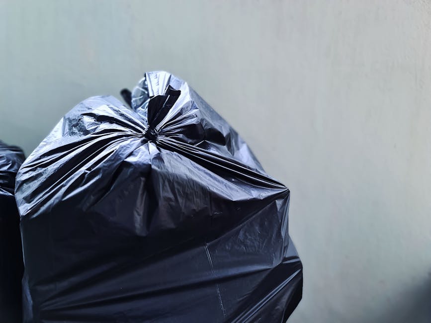 Close-up view of a tightly knotted black plastic rubbish bag with a shiny, slightly crinkled surface, positioned on the left side of the image against a plain, light-colored wall background. The bag is filled with waste and appears to be on a paved or concrete surface, possibly on a driveway or pavement. The lighting is even, highlighting the texture and creases in the plastic material. The scene suggests a setting where waste is prepared for collection, aligning with private rubbish removal services such as those offered by Rubbish Removal Islington, and illustrating the typical appearance of waste ready for alternative disposal methods outside local authority collections.