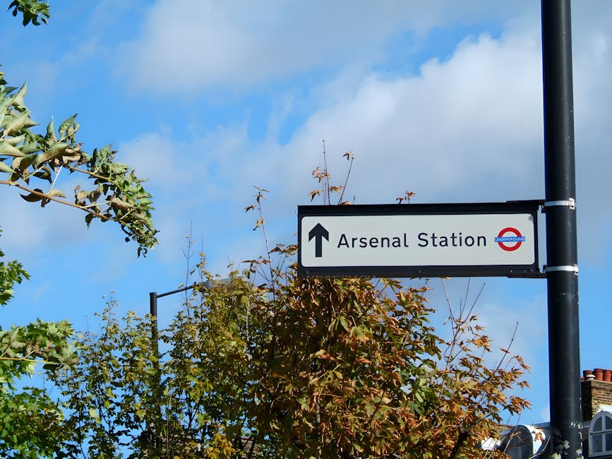 A rectangular white directional road sign mounted on a black metal pole points straight ahead, indicating the way to Arsenal Station. The sign features bold black text reading 'Arsenal Station' and includes the iconic red, blue, and white London Underground roundel logo on the right side. Surrounding the pole are leafy trees with green and brown foliage, some branches extending into the frame, suggesting a mild, overcast day with scattered clouds visible in the bright blue sky. The scene appears to be in an urban area with rooftops and chimney stacks visible in the background. The overall atmosphere is calm, with natural light illuminating the scene, and the sign situated near a pavement or street area, subtly associated with alternative directions for public transportation and local area navigation, as seen in the context of managing urban waste removal or related services by companies such as Rubbish Removal Islington.