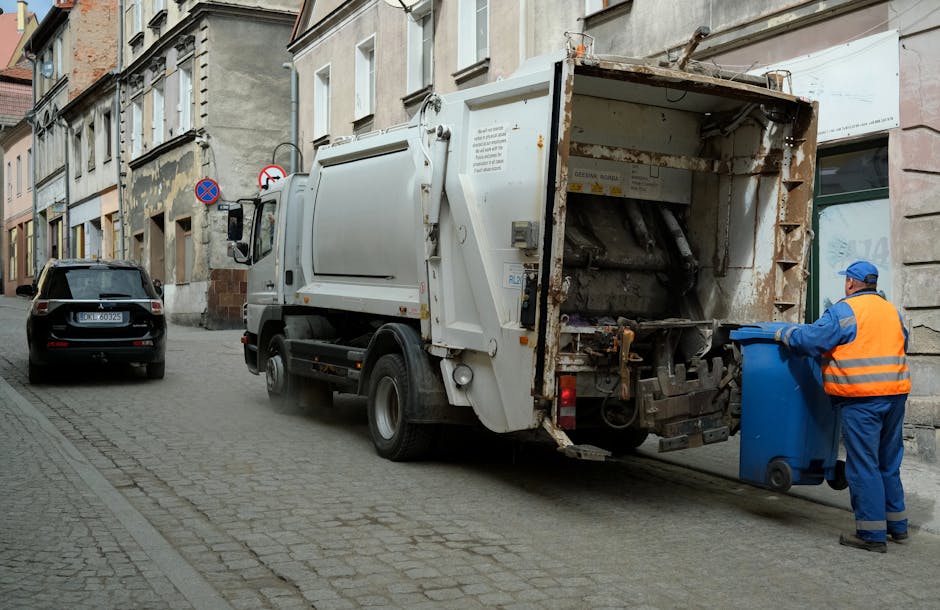 A commercial waste collection truck with a white metal body and a large open rear compactor, positioned on a narrow urban street with cobblestone paving. The truck's rear section shows visible rust and grime, with mechanical parts and hydraulic components exposed as it is in the process of emptying a blue wheeled refuse bin. A worker dressed in blue coveralls and a high-visibility orange vest is pushing the bin towards the truck's open rear compartment. To the left of the truck, a black SUV is parked alongside old, multi-story buildings with a mix of plaster and brick facades, some with peeling paint and weathered surfaces. Nearby, a 'no parking' traffic sign is attached to a pole. The scene is lit with neutral, natural daylight, emphasizing the utilitarian environment typical of independent waste collection services in urban areas, aligning with the themes of rubbish removal and private waste handling.