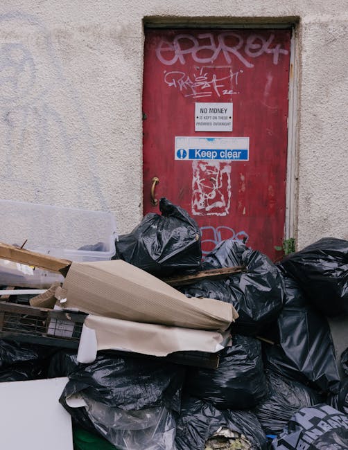 A pile of black rubbish bags, some partially torn or crumpled, stacked against a textured beige wall beneath a red metal door that features graffiti tags at the top, a white sign stating 'NO MONEY, NO CASH ON DELIVERY, PAYMENTS OVERDUE' in black text, and a blue and white sticker that reads 'Keep clear.' The rubbish bags are filled with household waste and are resting on a mix of cardboard and plastic packaging, with some items like a small section of white foam or polystyrene visible among the debris. The environment suggests an alleyway or service area where private waste collection or on-site clearance might be conducted, aligning with independent rubbish removal services typical of companies like Rubbish Removal Islington. The scene is illuminated by natural daylight, casting subtle shadows on the ground and emphasizing the texture of the wall and the waste materials, reflecting a typical rubbish disposal situation encountered during non-standard waste handling scenarios in a residential or commercial setting.