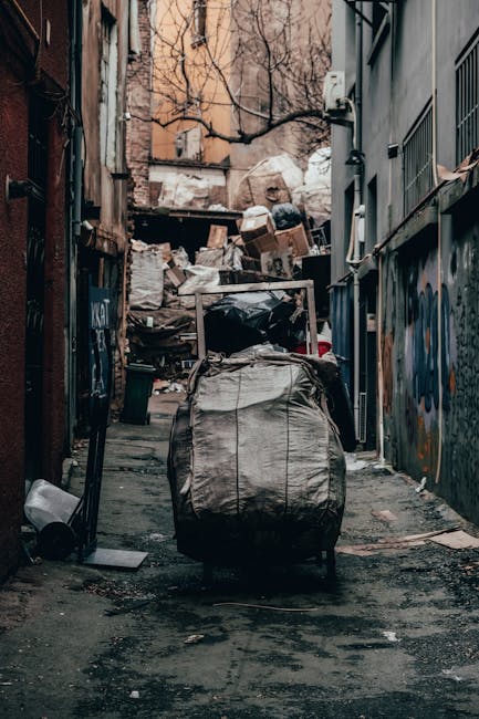 A narrow urban alleyway filled with overflowing piles of mixed waste, including cardboard boxes, plastic bags, and other refuse stacked against a back wall and spilling onto the ground. In the foreground, a large, grey, industrial-style rubbish bag is positioned on a small wheeled trolley, ready for collection or disposal. The surrounding environment features dark, weathered brick walls on the left side and a grey metal fence with barred windows on the right, both adorned with graffiti. Bare tree branches extend overhead, casting minimal light into the scene, which appears cluttered and unmaintained. The background showcases piled waste, partially obstructing a narrow access point with a makeshift barrier, highlighting the need for efficient rubbish removal. This scene exemplifies an unmanaged alley awaiting private waste collection, such as services provided by Rubbish Removal Islington, illustrating the importance of timely and professional rubbish clearance to maintain urban cleanliness.