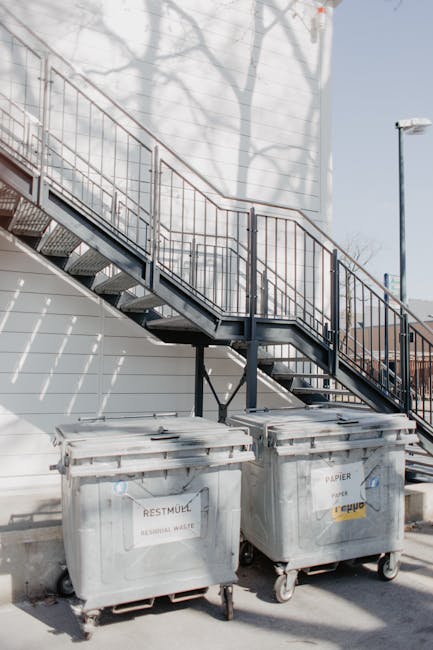 The image depicts two large grey wheeled waste containers positioned under an outdoor metal staircase with a railing, against the side of a building with horizontal siding painted in a light color. The containers are marked with labels indicating they are for different types of waste; one primarily for residual waste and the other for paper, with the label on the paper container featuring a yellow and white design and the residual waste container labeled 'RESTMÜL' in German. The containers are made of sturdy plastic, showing some signs of use with minor scuffs, and are situated on a paved surface, likely a street or alleyway. The staircase above them is constructed from metal, painted dark gray or black, and features grated steps, with shadows of nearby tree branches cast onto the wall behind. A street lamp and some additional buildings or structures are visible in the background, suggesting an urban environment. This scene relates to waste management and rubbish disposal services, aligned with private or alleyway collection options often employed by organisations like Rubbish Removal Islington, especially in contexts outside of standard council waste collection routes.