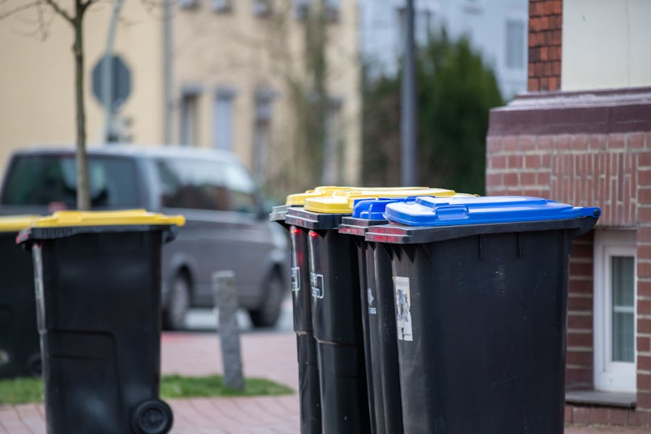 A row of three large black wheelie bins with plastic lids, positioned on a paved sidewalk adjacent to a red brick building. The lids of the bins are colored yellow, blue, and black, from left to right, with the blue lid slightly ajar on the rightmost bin. The surface of the bins appears smooth and slightly glossy, reflecting ambient light. In the background, there is a parked grey van on the street, partially obscured by the bins, with a residential area featuring trees, light-colored buildings, and utility poles visible further back. The scene is captured during daylight, with natural lighting highlighting the textures of the plastic bins, brickwork, and surroundings. This image exemplifies typical kerbside waste collection setups, relevant to private rubbish disposal options provided by services such as Rubbish Removal Islington, aligned with understanding collection practices and waste management regulations in the area.