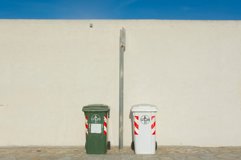 The image depicts two large grey wheeled waste containers positioned under an outdoor metal staircase with a railing, against the side of a building with horizontal siding painted in a light color. The containers are marked with labels indicating they are for different types of waste; one primarily for residual waste and the other for paper, with the label on the paper container featuring a yellow and white design and the residual waste container labeled 'RESTMÜL' in German. The containers are made of sturdy plastic, showing some signs of use with minor scuffs, and are situated on a paved surface, likely a street or alleyway. The staircase above them is constructed from metal, painted dark gray or black, and features grated steps, with shadows of nearby tree branches cast onto the wall behind. A street lamp and some additional buildings or structures are visible in the background, suggesting an urban environment. This scene relates to waste management and rubbish disposal services, aligned with private or alleyway collection options often employed by organisations like Rubbish Removal Islington, especially in contexts outside of standard council waste collection routes.