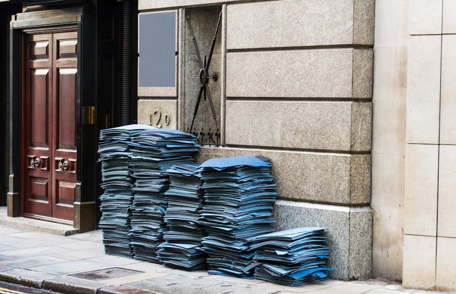 A large stack of blue and black plastic folders is piled up on the pavement outside a building with a stone exterior wall. The folders appear crumpled and are arranged in uneven towers of varying heights, some leaning slightly. To the left, there is a wooden double door with a dark finish, partially obscured by the piles. Behind the folders, a metal security shutter is rolled up, revealing a rectangular intercom or utility box attached to the stone wall. The scene is illuminated by natural daylight, casting soft shadows on the textured surface of the folders and the building façade. The environment suggests an area adjacent to a commercial or residential building where private waste collection or on-site clearance may be taking place, and this image is representative of services like rubbish removal provided by companies such as Rubbish Removal Islington, highlighting the need for proper disposal of accumulated paperwork or lightweight waste materials.