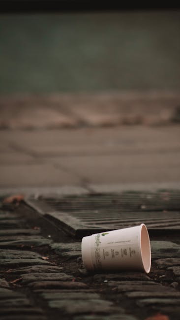 A discarded white paper cup with green text lies on a cobbled pavement next to a black metal drain cover. The cup appears crumpled and is positioned on its side, with part of it touching the edge of the drain. The surrounding area includes worn, uneven cobblestones with visible dirt and debris, and a blurred background suggests an outdoor environment, possibly near a building or sidewalk in Islington. This scene illustrates typical urban rubbish that Rubbish Removal Islington might handle as part of an on-site clearance or private waste collection service, emphasizing the everyday presence of waste in public outdoor spaces.