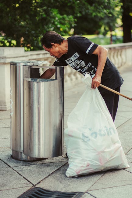 A middle-aged woman with short dark hair, dressed in a black t-shirt with white text and black trousers, is seen placing a large white plastic bag filled with rubbish into a stainless steel litter bin on a paved sidewalk. She is slightly bent over, holding the bag with her left hand while using her right hand to reach into the bin’s opening. The bin is part of a set of three cylindrical, polished metal containers positioned alongside each other, designed for waste collection. The background features lush green foliage and trees, indicating an outdoor urban environment, with a low stone wall or balustrade nearby. The lighting suggests daytime, with natural sunlight illuminating the scene. The woman appears to be engaged in a street cleaning or waste disposal activity, which is relevant to waste management services such as those offered by Rubbish Removal Islington, especially in the context of providing independent waste clearance options for local communities and businesses.