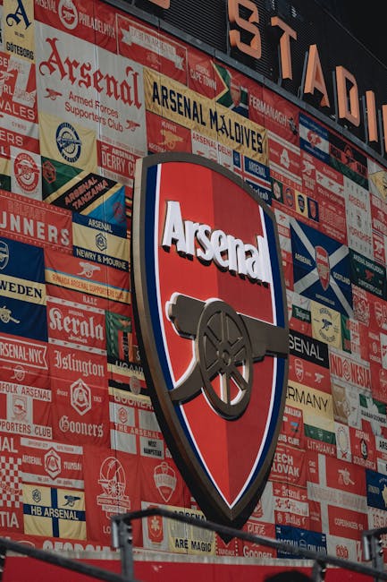 A large outdoor wall display featuring numerous football club banners and crests, predominantly in red, white, and blue colors, with some gold and black accents. The Centenary Arsenal crest, centered in the image, includes a shield with the word 'Arsenal' in white lettering on a red background, topped by a curved black cannon with a metallic finish. The display appears to be mounted on a building or stadium exterior, with the banners arranged in a collage-like manner, creating a visually dense background. The lighting is bright and natural, casting minimal shadows. The overall scene emphasizes the Arsenal football club's branding and supporter memorabilia, capturing the vibrant atmosphere of the club's heritage, which can be relevant in the context of alternative ways fans or clubs display and manage their memorabilia outside of standard storage or waste disposal. Rubbish Removal Islington may occasionally handle waste related to such memorabilia when clearing fan areas or storage spaces, though this image primarily illustrates the collection and display of club-branded items.