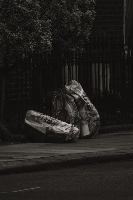 Two large black plastic rubbish bags filled with waste are placed on a paved sidewalk in front of a black metal fence and dense foliage of trees or bushes. The bags appear bulging and are tightly sealed, indicating they contain household or general waste. The scene is set outdoors on a street with a curb, and there is minimal natural light, giving the image a somewhat subdued or overcast appearance. The bags are positioned close to the fence, which has vertical bars and a section that appears to be a gate, suggesting a residential or private property boundary. The environment reflects an informal or private rubbish disposal scene, potentially requiring collection or removal services, as seen with companies like Rubbish Removal Islington. Overall, the image emphasizes typical curbside waste awaiting collection, highlighting the importance of appropriate disposal and clearance processes for domestic or commercial refuse.